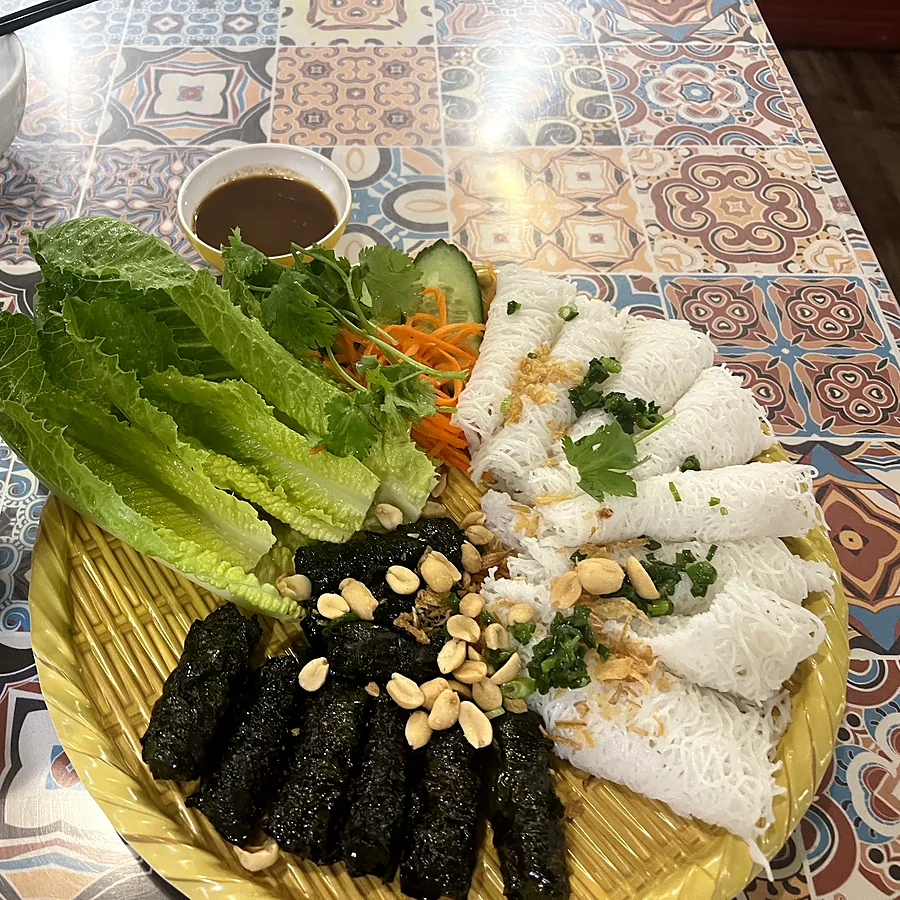 Grilled beef in betel leaves with woven rice vermicelli, lettuce, peanuts and dipping sauce on a patterned tile table